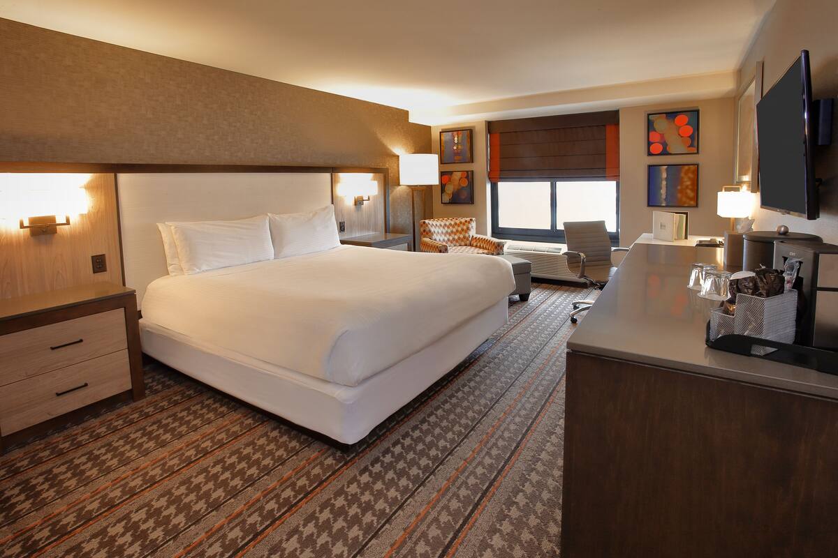 A hotel room with a large bed, a desk with a flat-screen TV, a chair, and a desk lamp.