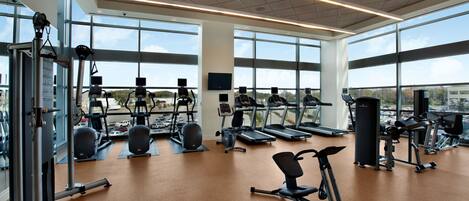 Un gimnasio espacioso con diversos equipos de ejercicio, ventanas amplias y un techo alto.