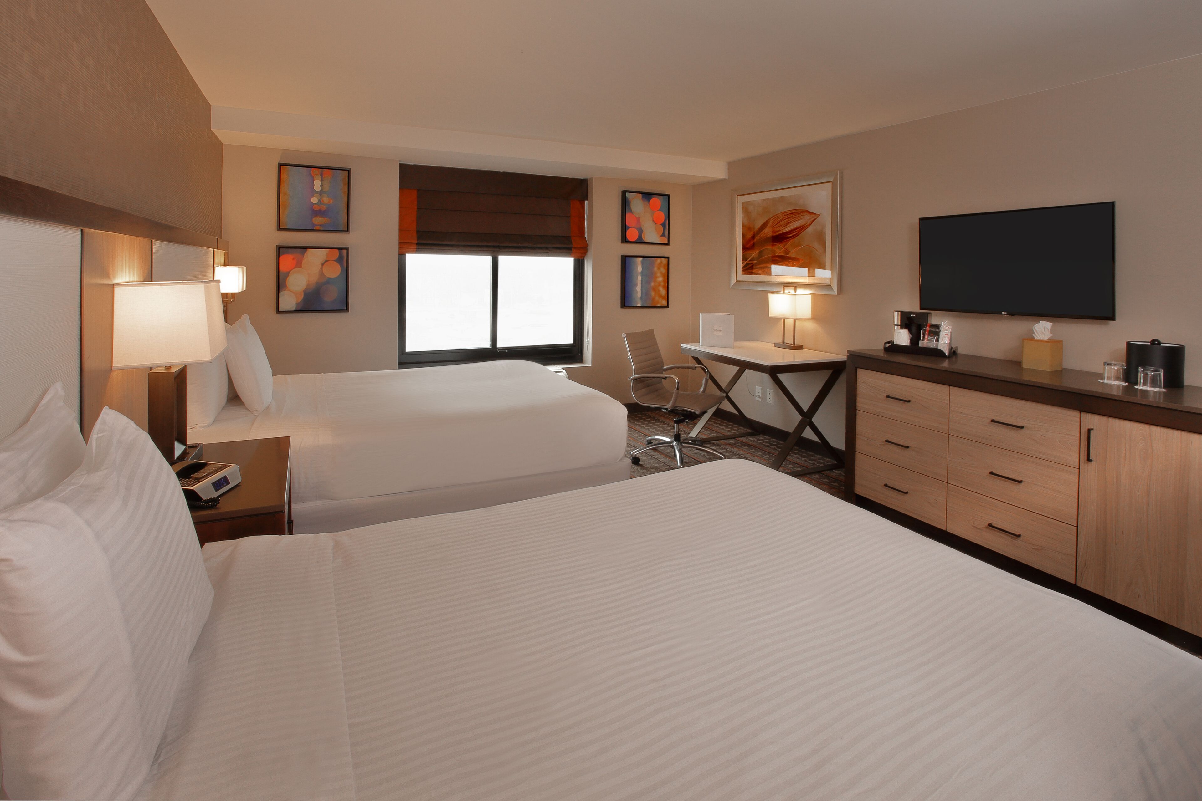 A hotel room with two beds, a desk, a chair, a TV, and artwork on the walls.