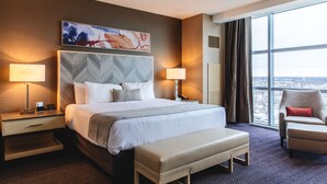 Een hotelkamer met een groot bed, twee nachtkastjes, een stoel, een bankje en een groot raam met uitzicht op de stad.