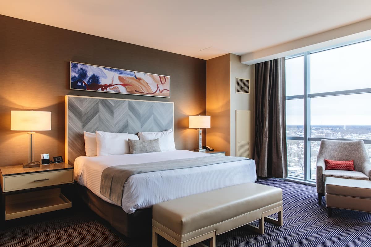 A hotel room with a large bed, two bedside tables, a chair, a bench, and a large window offering a city view.
