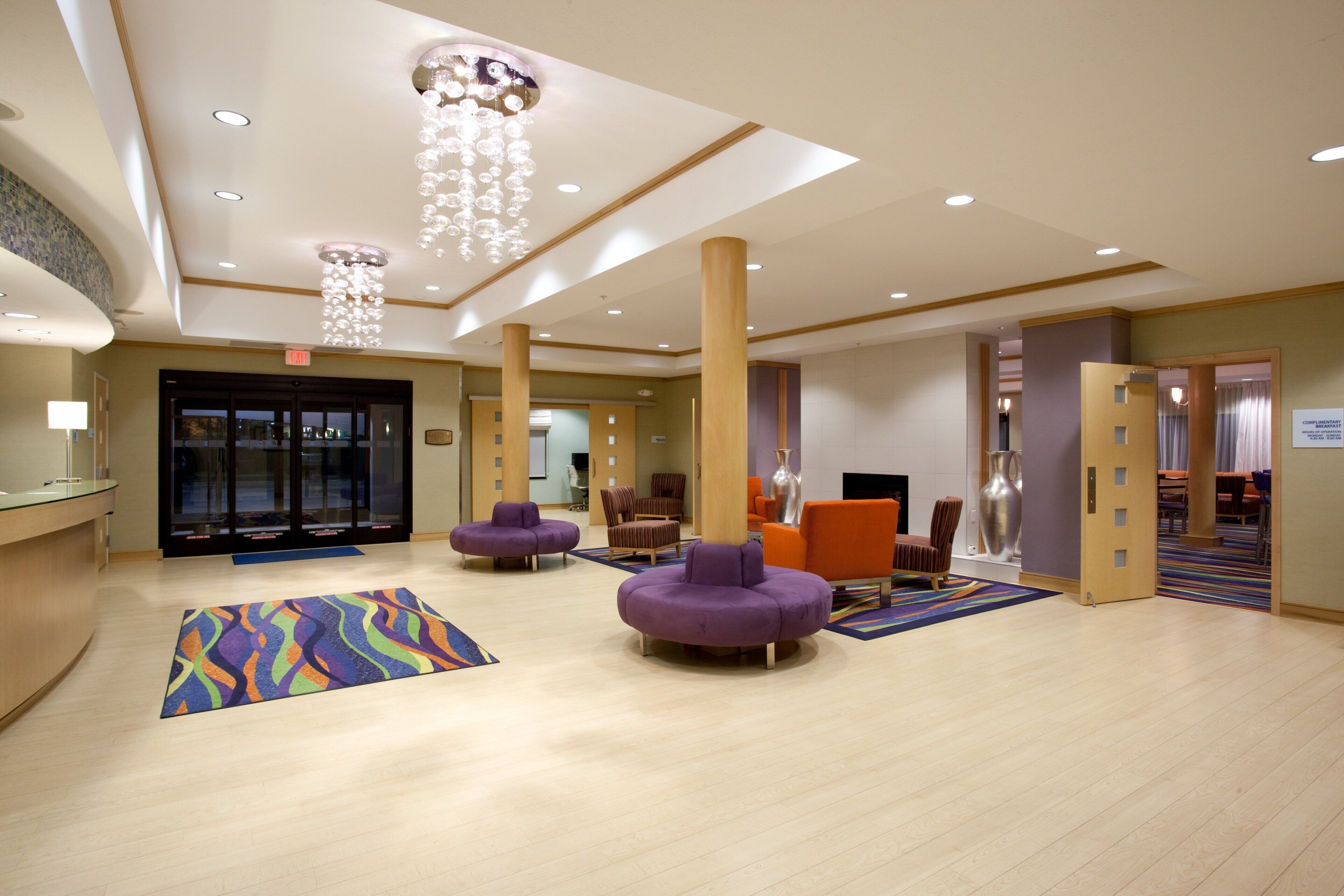 A hotel lobby with a reception desk, purple seating, a colorful rug, and a chandelier.