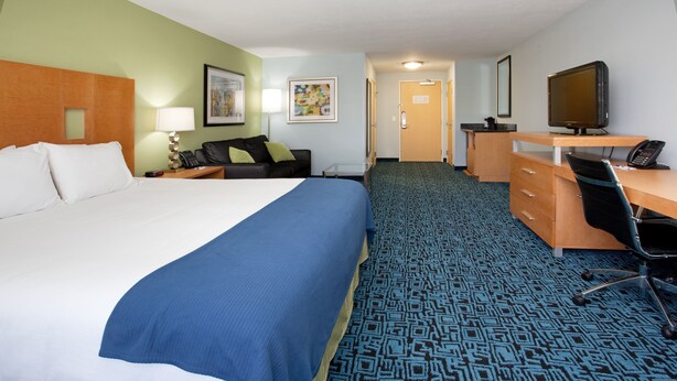 A hotel room with a large bed, a desk with a computer, a chair, a lamp, and a door leading to a bathroom.