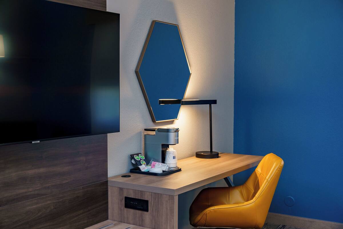 A hotel room with a wooden desk, a yellow chair, a coffee machine, a flat-screen TV, and a mirror on the wall.