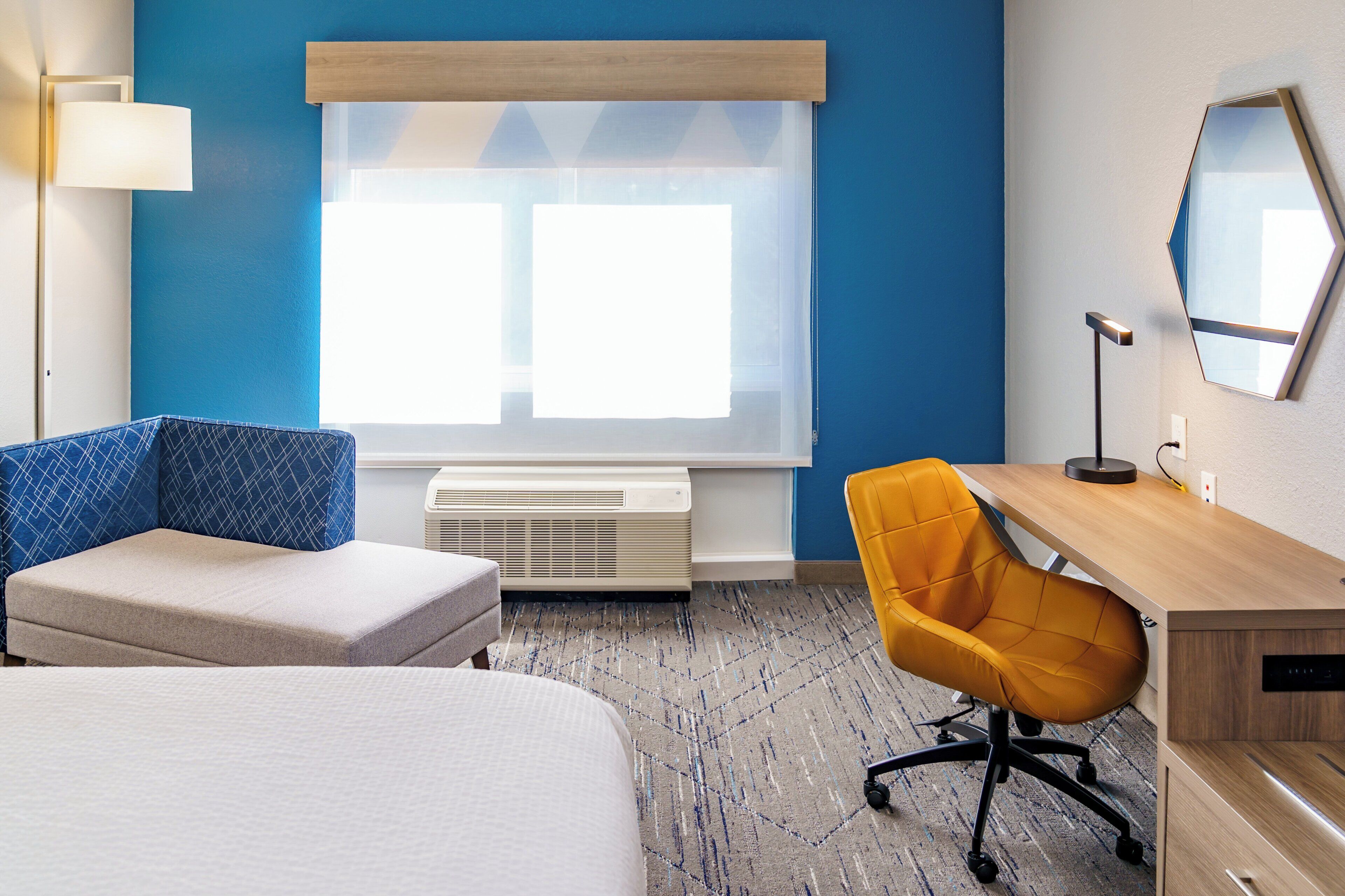Habitación de hotel con cama, escritorio, una silla naranja, lámpara y un ventanal con persianas.