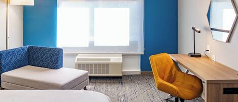 Une chambre d’hôtel avec un lit, un bureau, une chaise orange, une lampe et une grande fenêtre avec des stores.