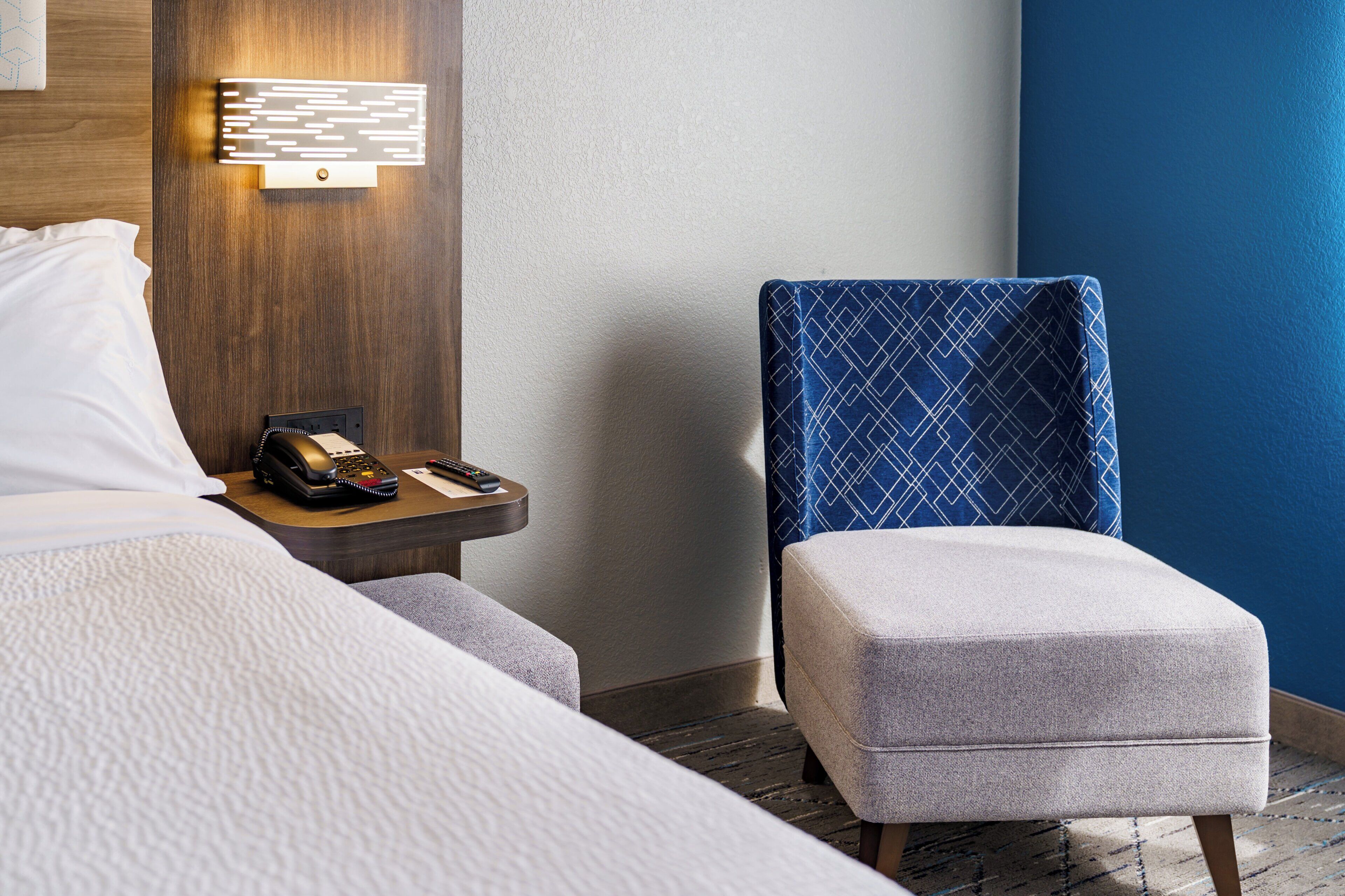 A hotel room with a bed, a nightstand with a phone and remote controls, a blue patterned chair, and a wall-mounted light fixture.