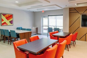 A modern dining area with wooden tables and orange chairs, a large window with a view, and a colorful abstract painting on the wall.