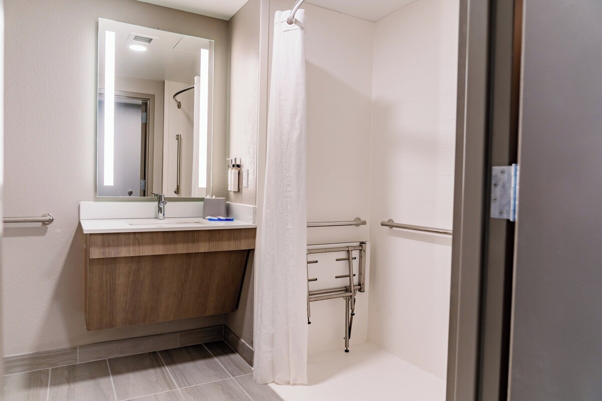 A modern bathroom with a wooden vanity, a large mirror, a shower with a curtain, and a grab bar.