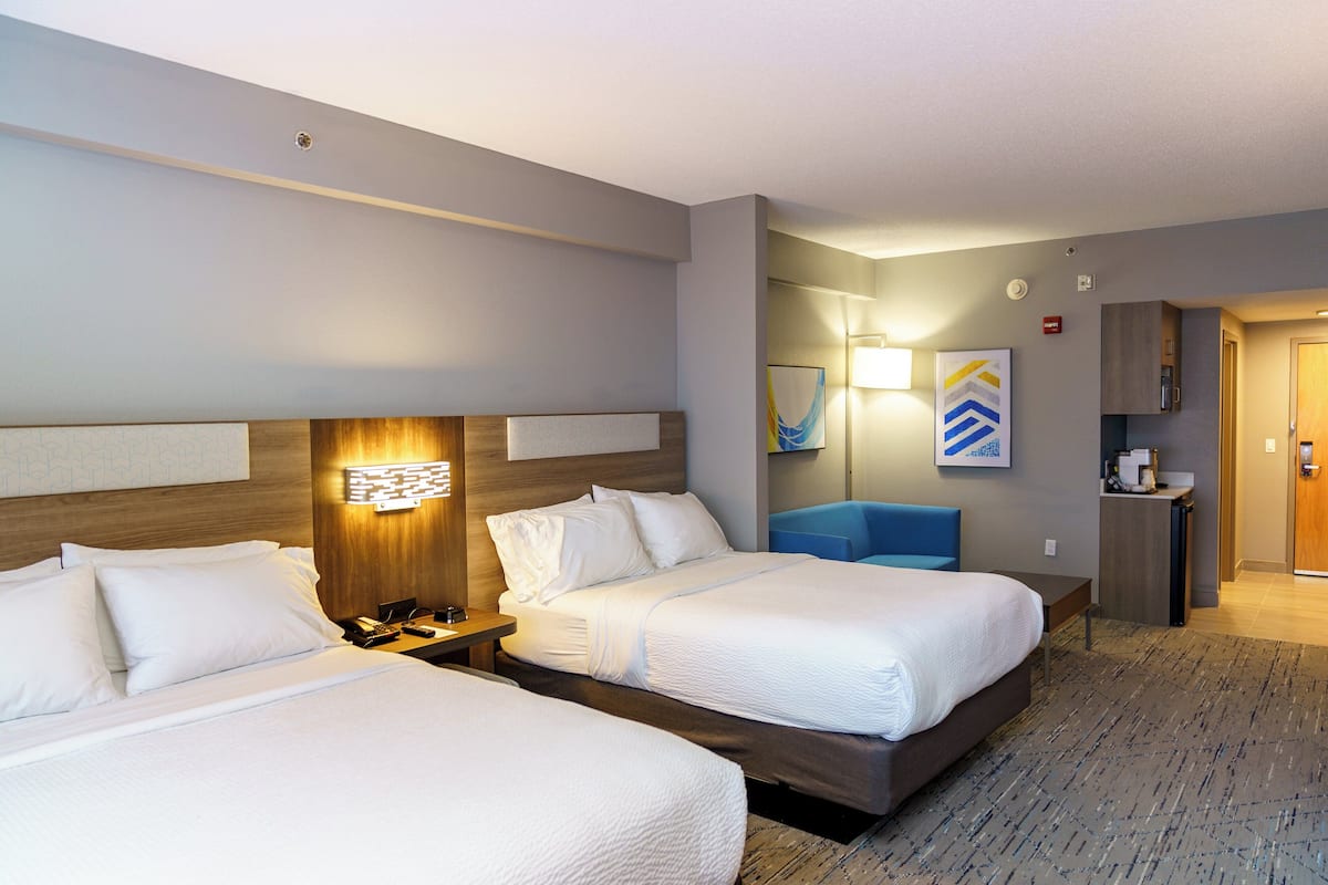 A hotel room with two beds, a desk, a lamp, a mirror, and a small kitchenette.