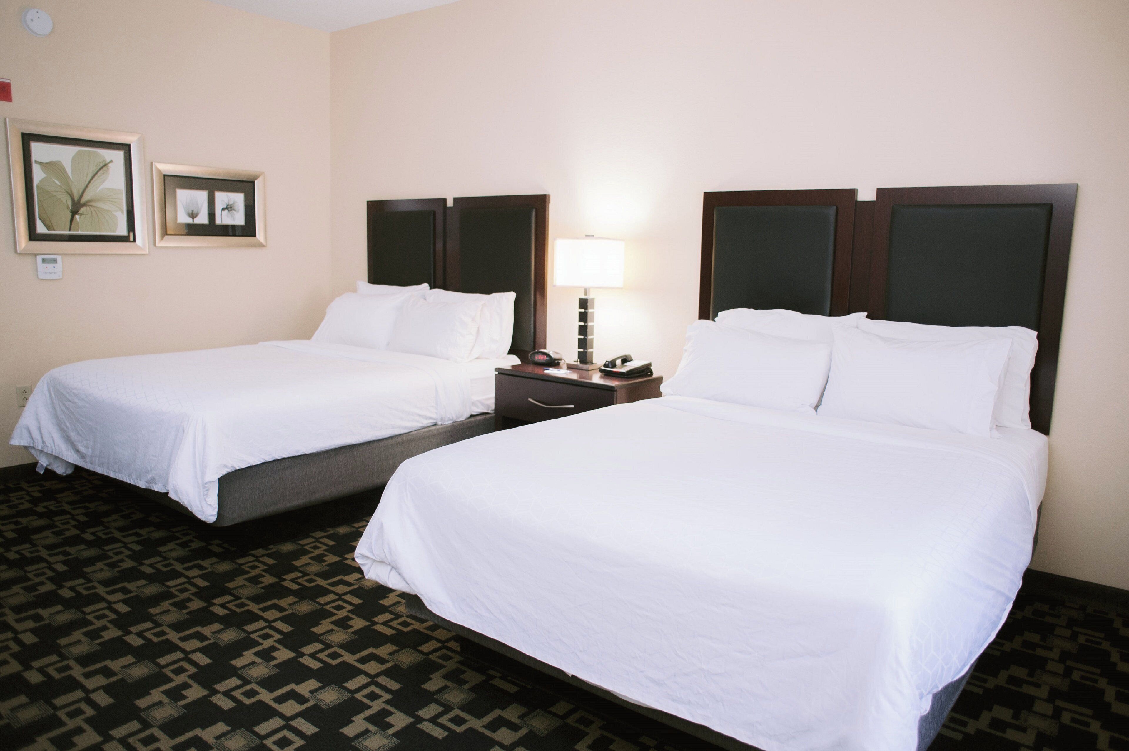 A hotel room with two single beds, a nightstand with a lamp, and a wall-mounted thermostat.