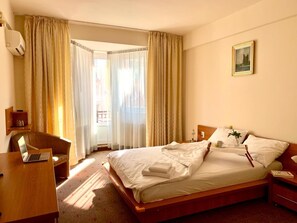 A hotel room with a bed, a desk with a laptop, a chair, a nightstand, and a painting on the wall. - Victoria Hotel Cluj Napoca (Cluj-Napoca)