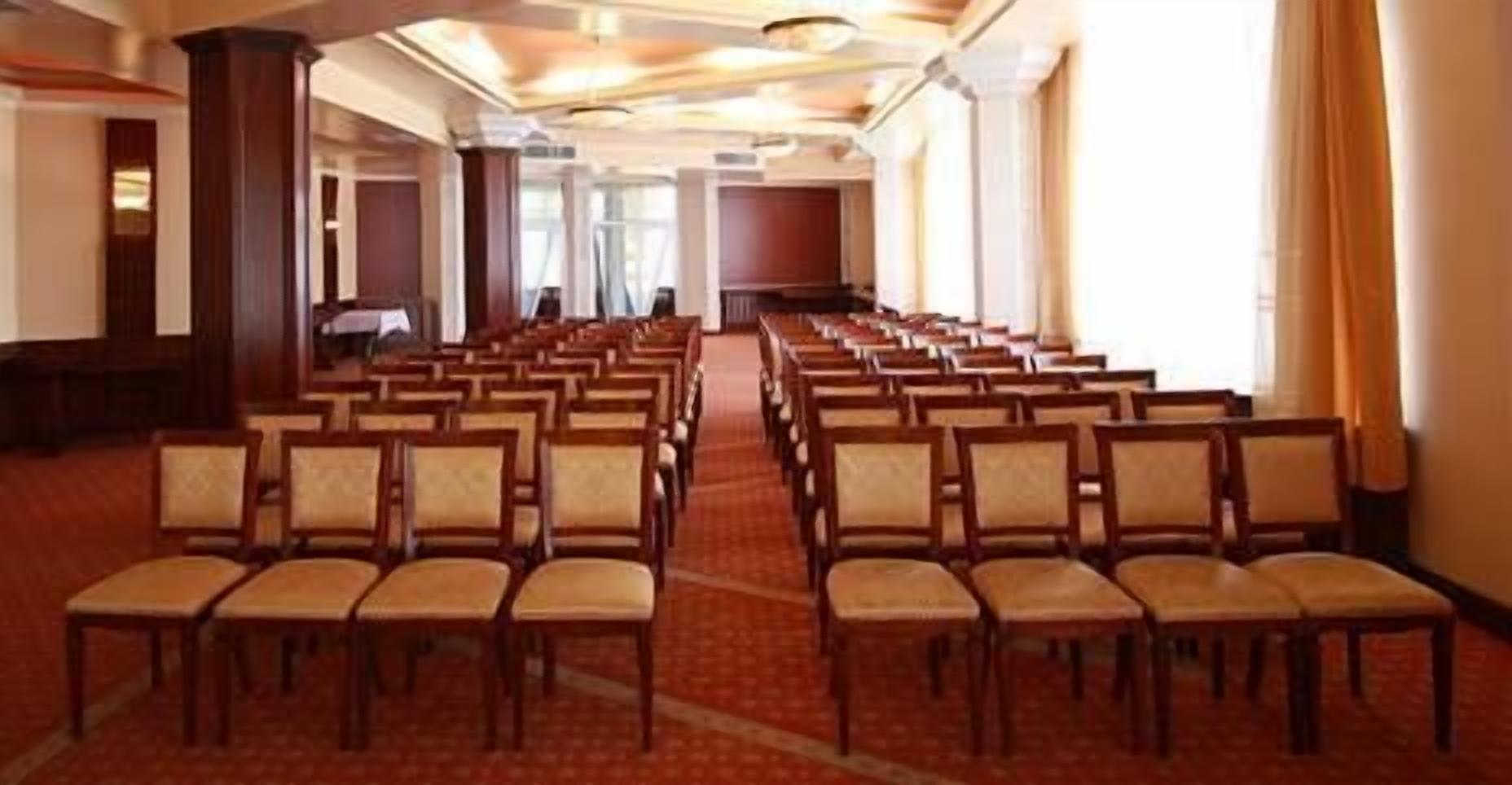 A conference room with rows of wooden chairs and tables, carpeted floor, and large windows with curtains.