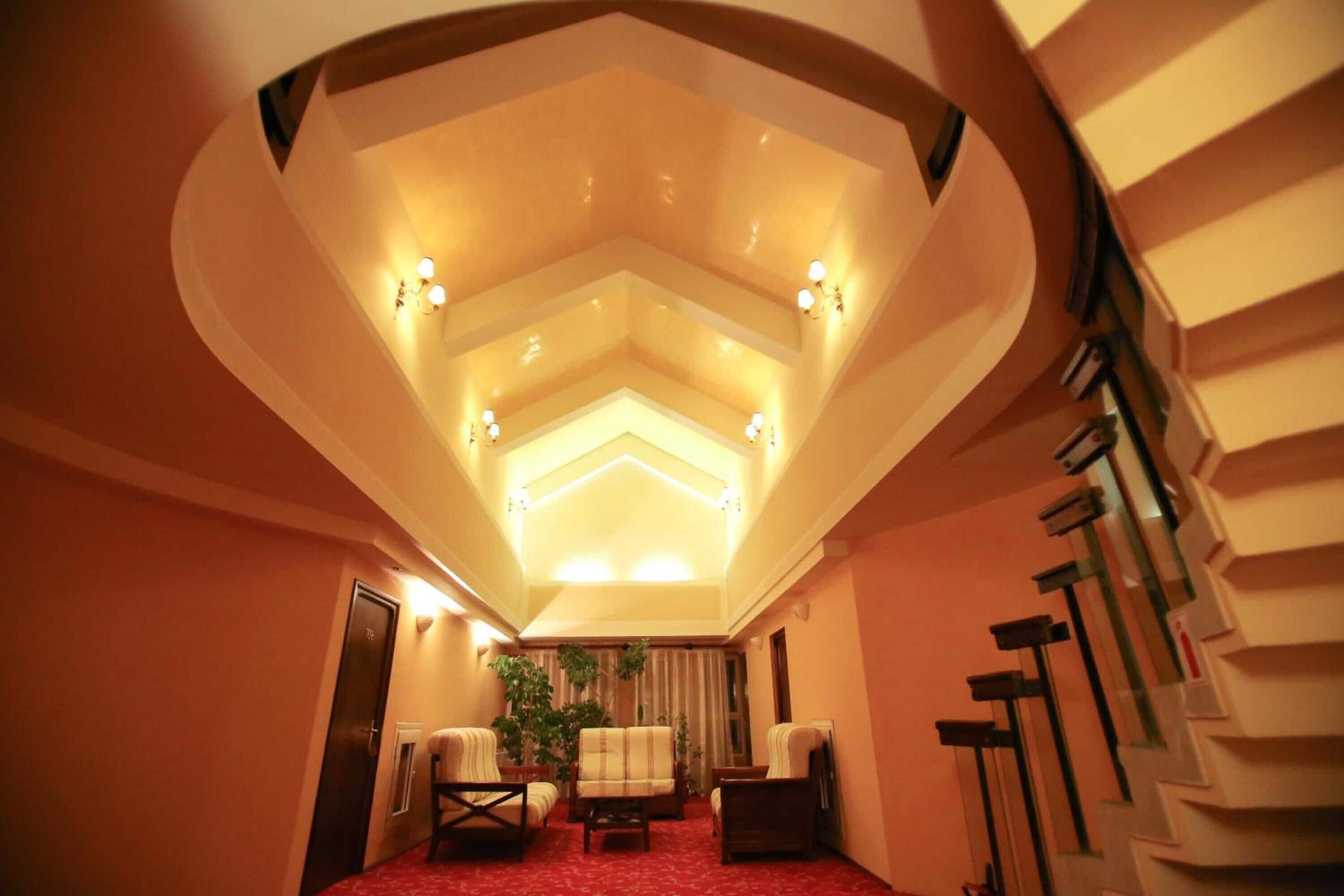 Habitación de hotel con techo alto, alfombra roja, zona de estar con sillas y una mesa, y un gran ventanal con cortinas.