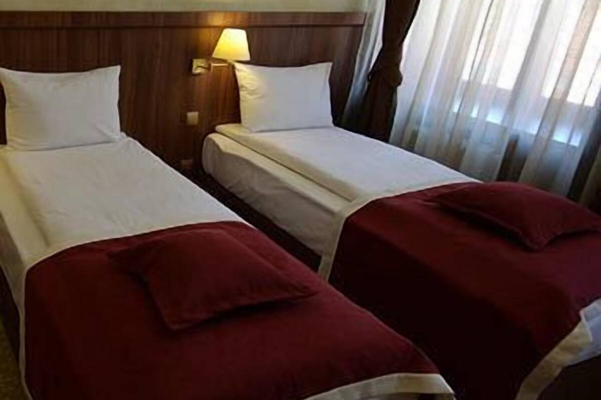 A hotel room with two beds, each with a white bedspread and red pillows. There is a nightstand with a lamp between the beds.