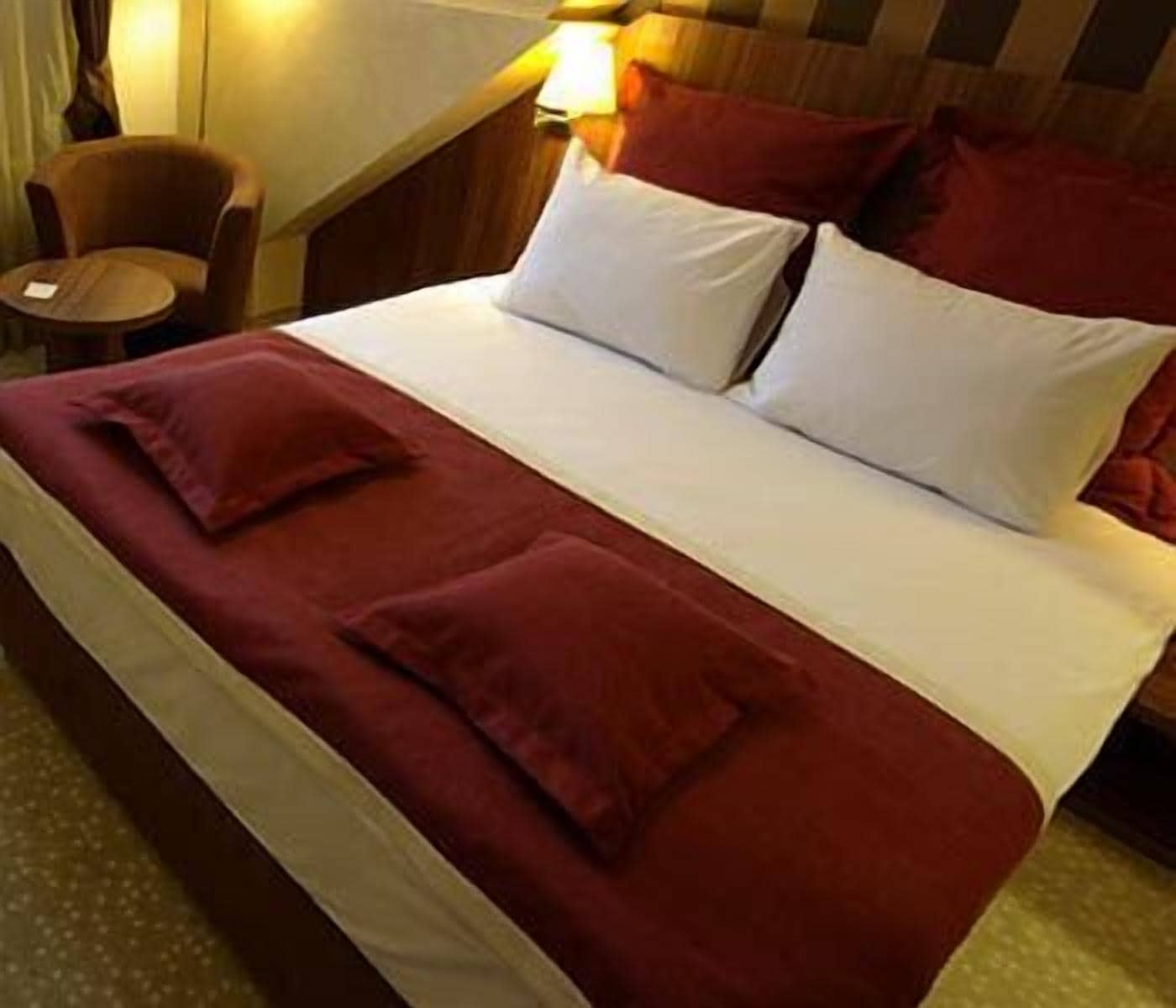Een bed met een rode deken en kussens, een klein rond tafeltje met een lamp, en een stoel.
