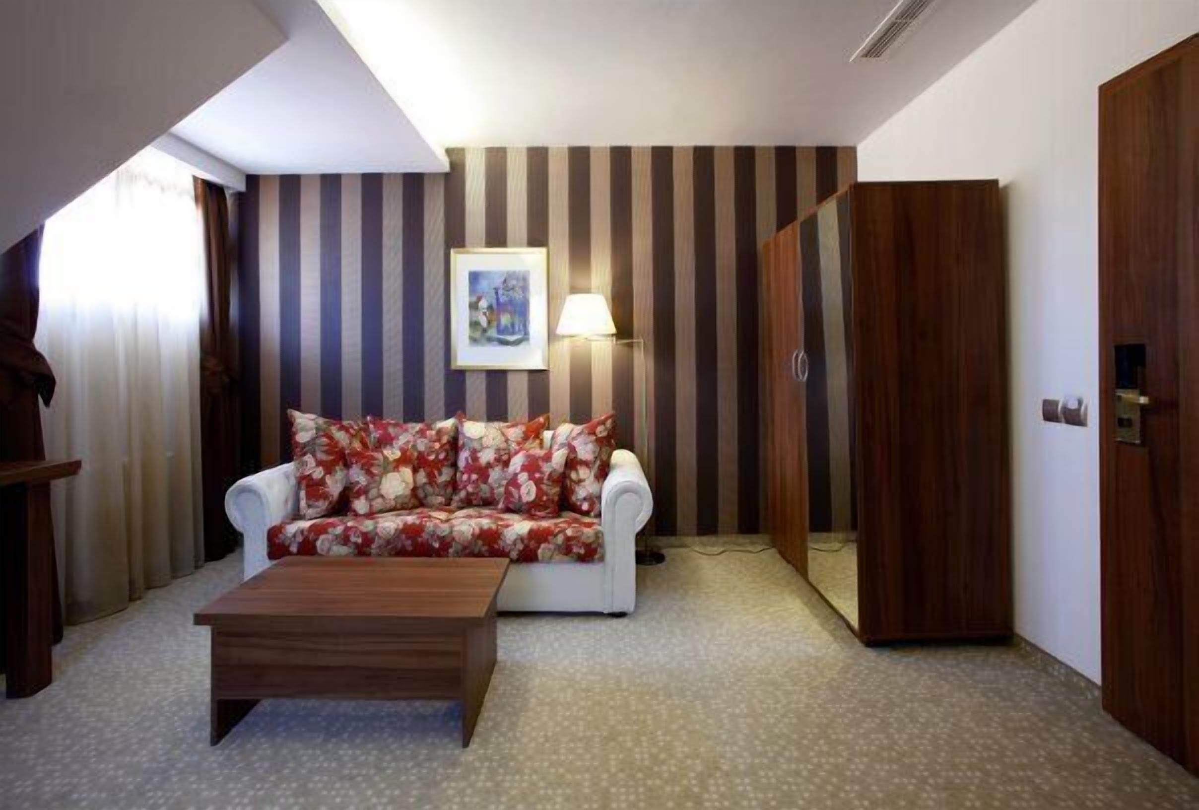 A room with a striped wall, a white sofa with floral cushions, a wooden coffee table, a lamp, and a wardrobe.