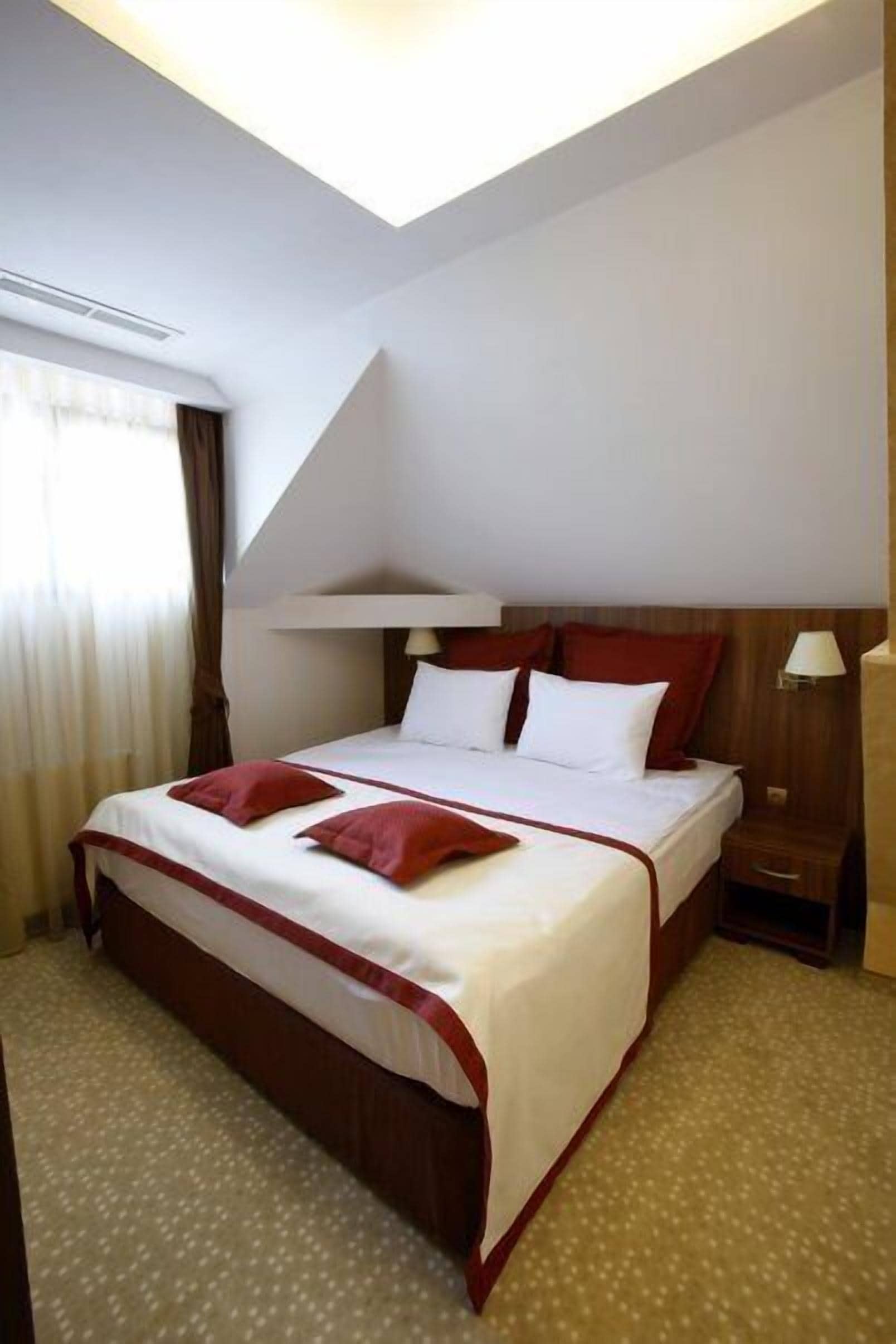 A hotel room with a large bed, a wooden headboard, a bedside table with a lamp, and a window with curtains.