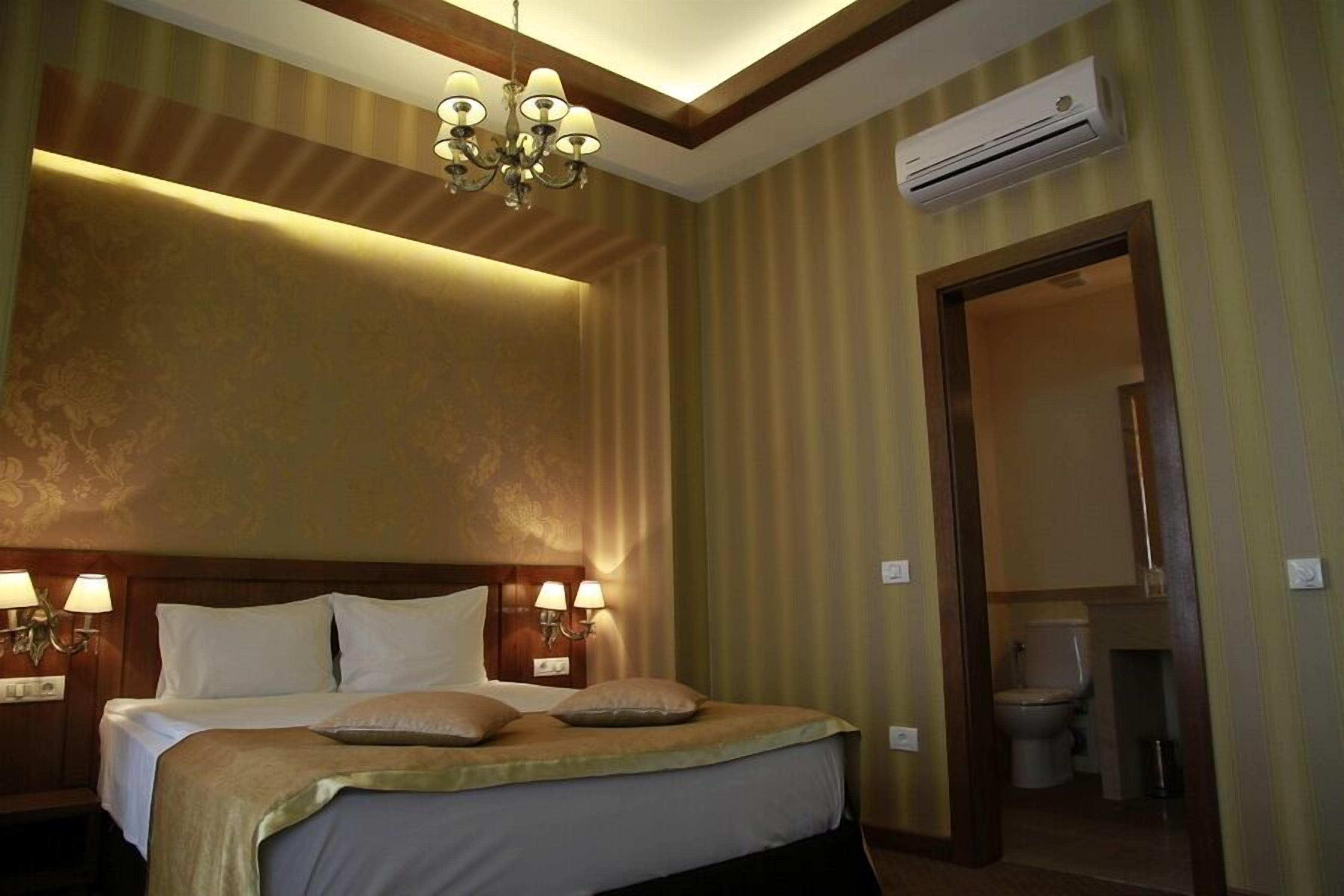 Habitación de hotel con una cama grande, dos lámparas de noche, un candelabro, un equipo de aire acondicionado y un baño a la vista.