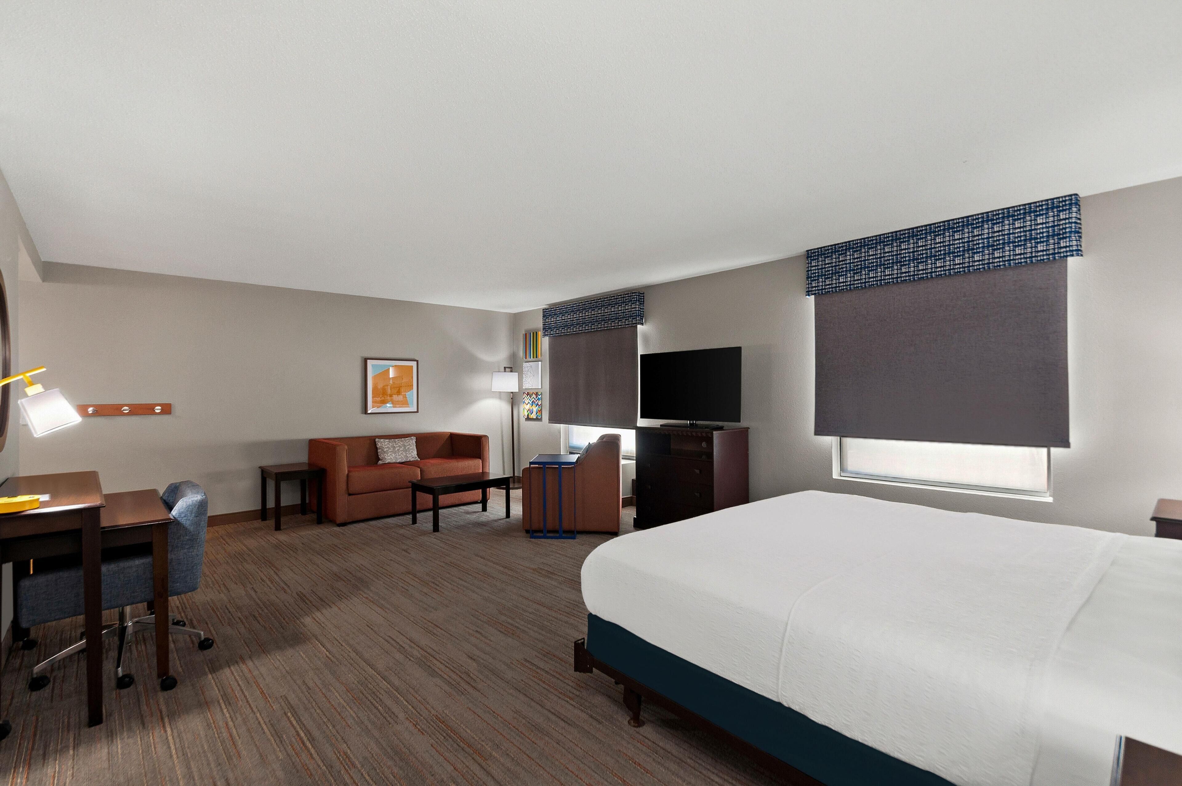 A hotel room with a bed, a desk, a sofa, a TV, and a window with blinds.