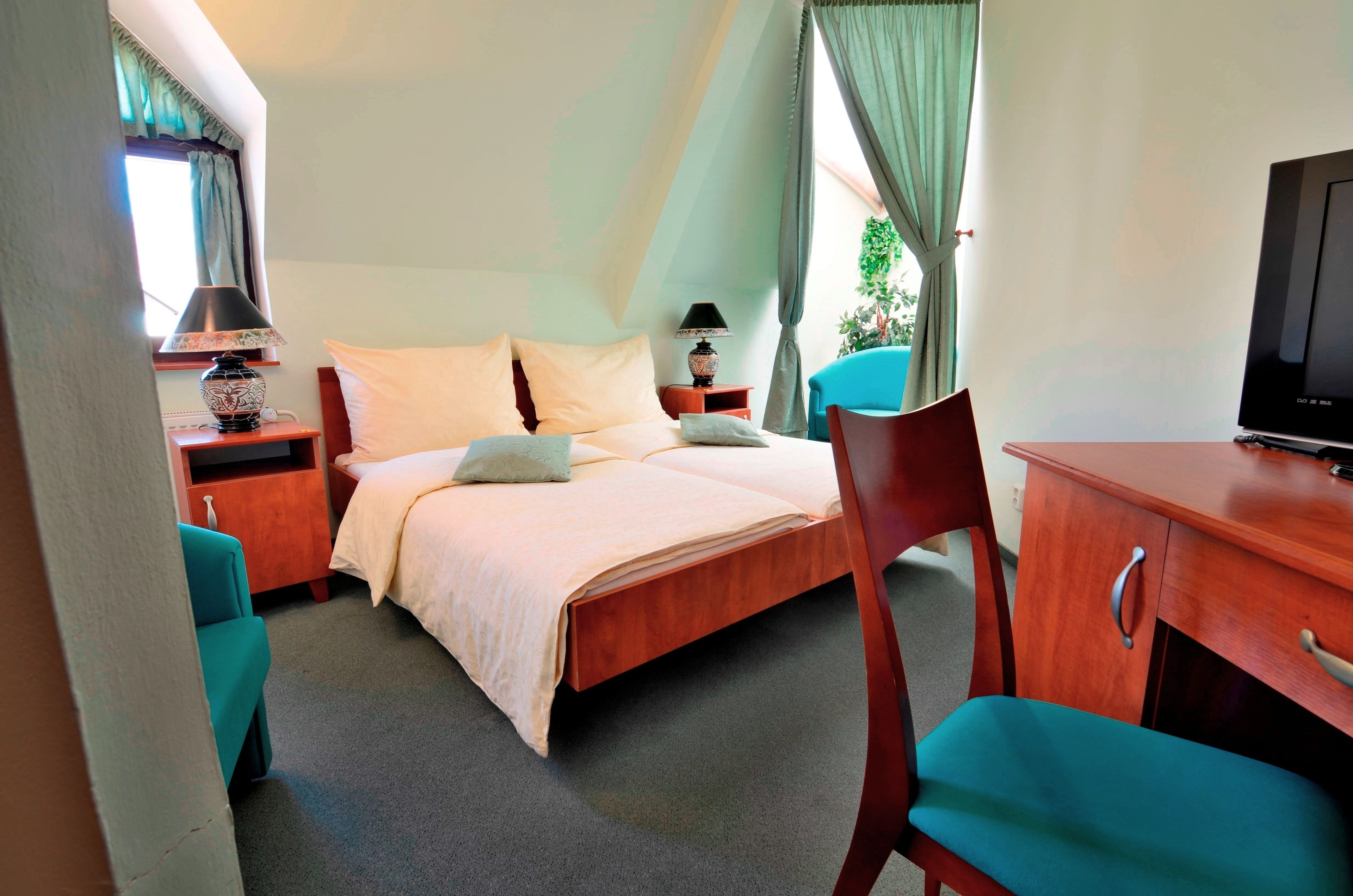 A hotel room with a bed, two bedside tables, a desk, a chair, a television, and a window with curtains.