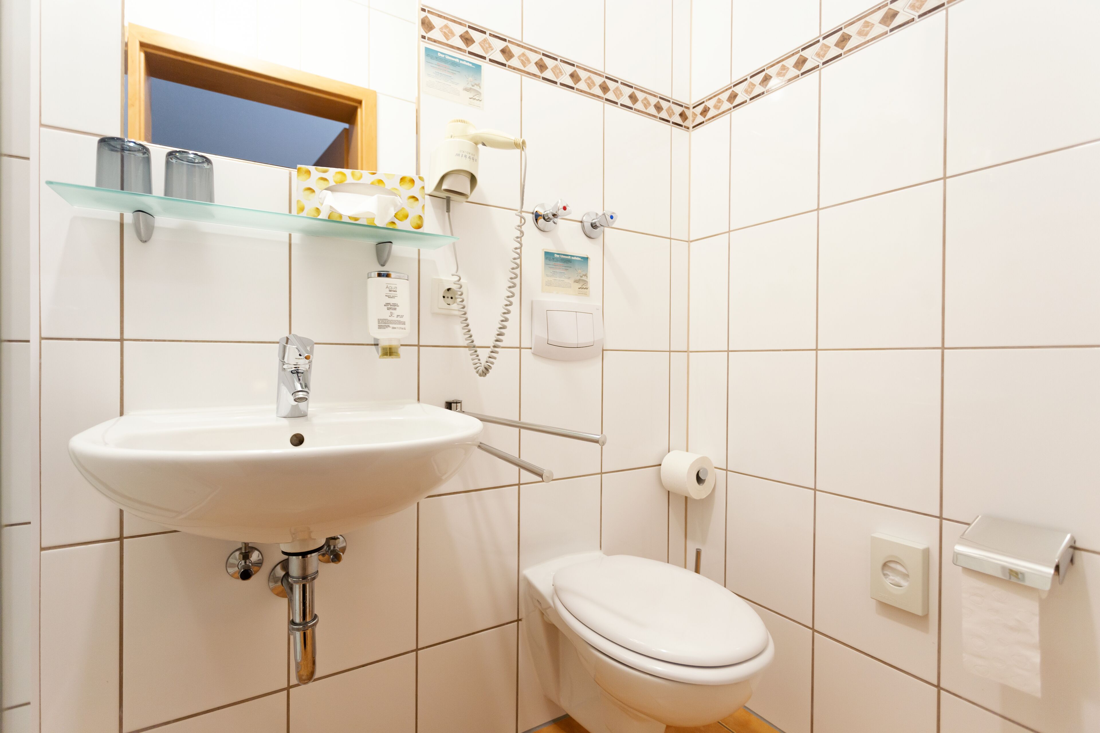 Un baño con un lavamanos blanco, un inodoro y un estante de vidrio sobre el lavamanos.