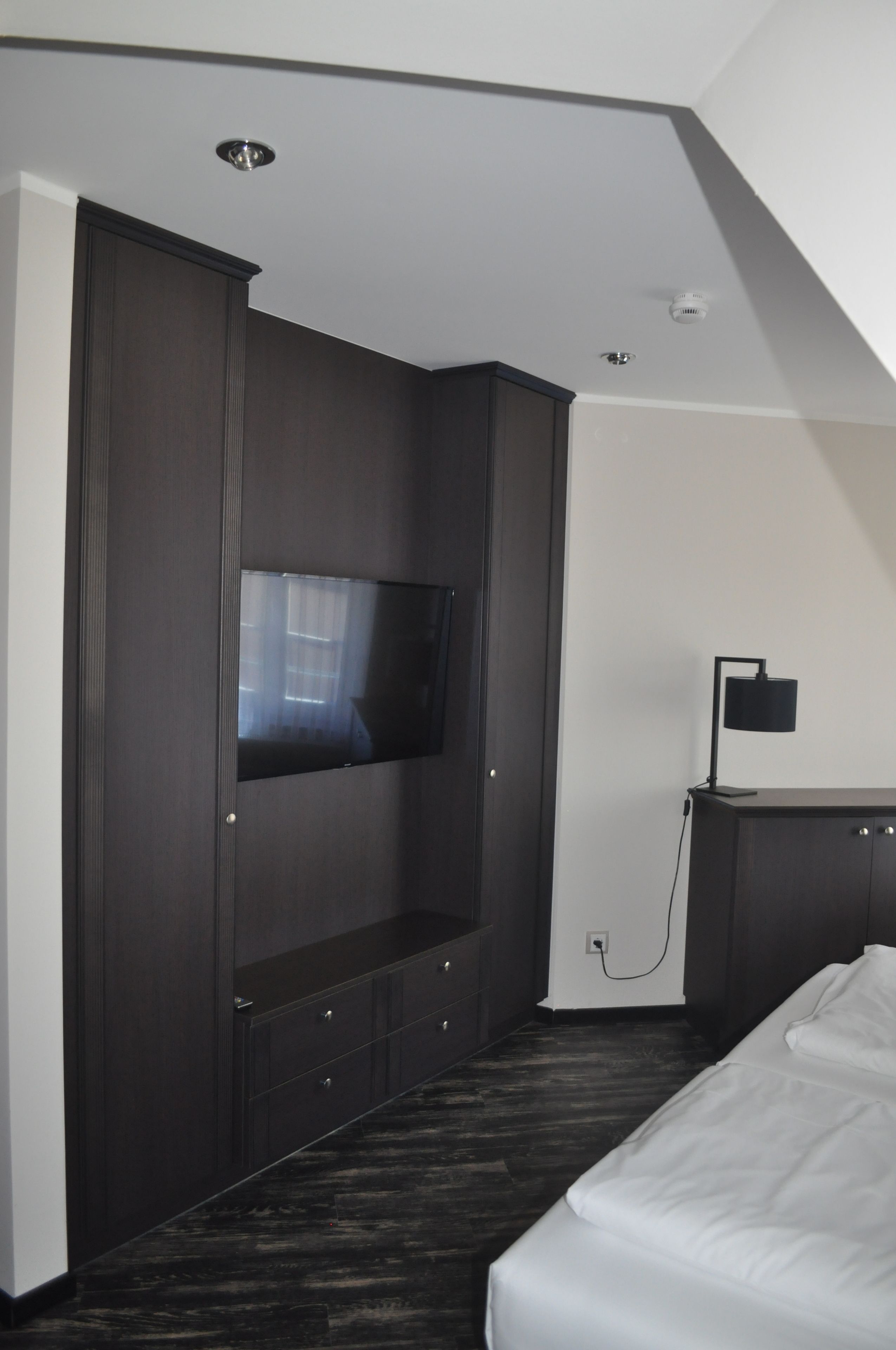 A hotel room with a dark wood wardrobe, a flat-screen TV mounted on the wall, a bedside table with a lamp, and a bed with white bedding.