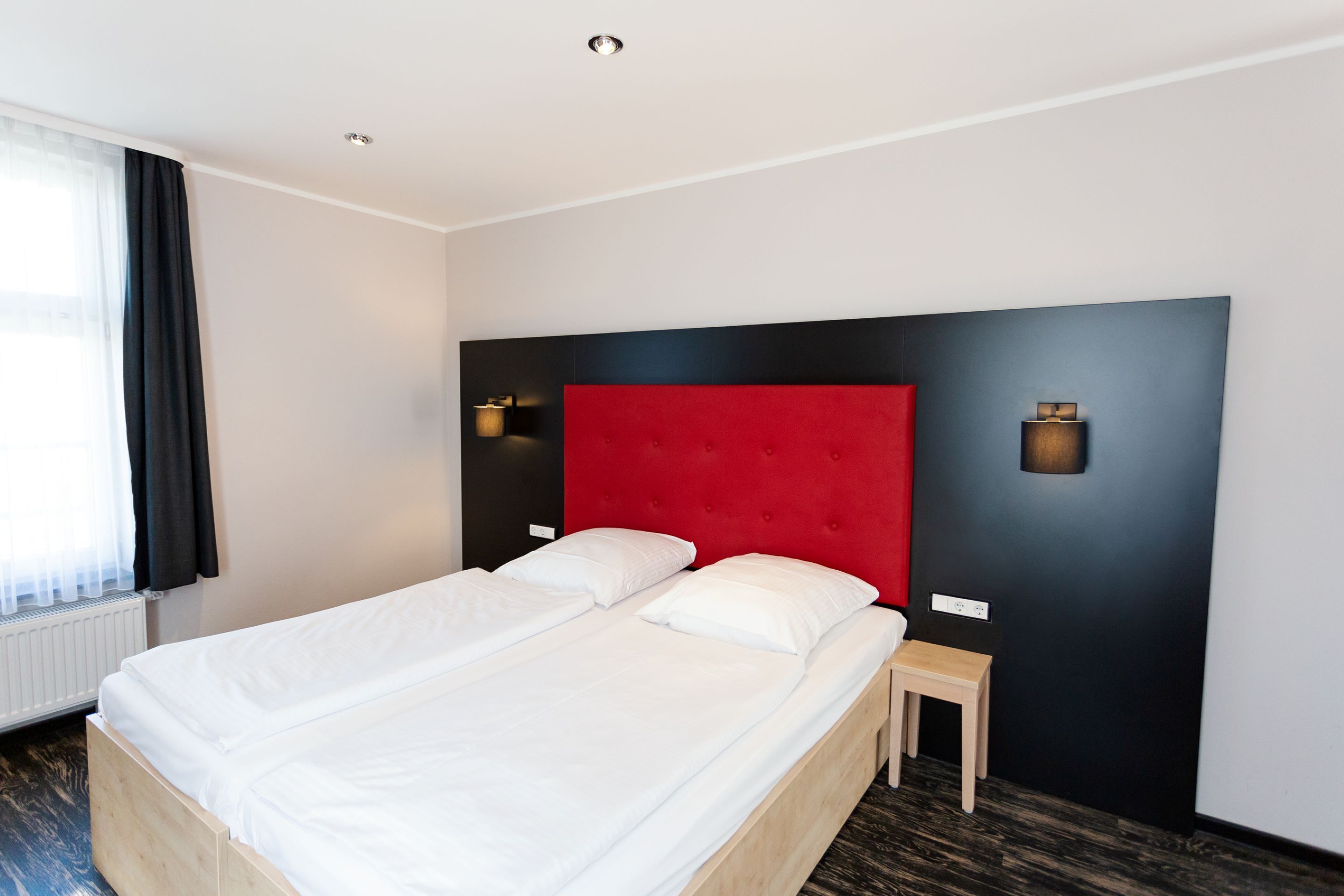 Moderni hotellihuone, jossa on suuri sänky, punainen pääty ja kaksi yöpöytävalaisinta.