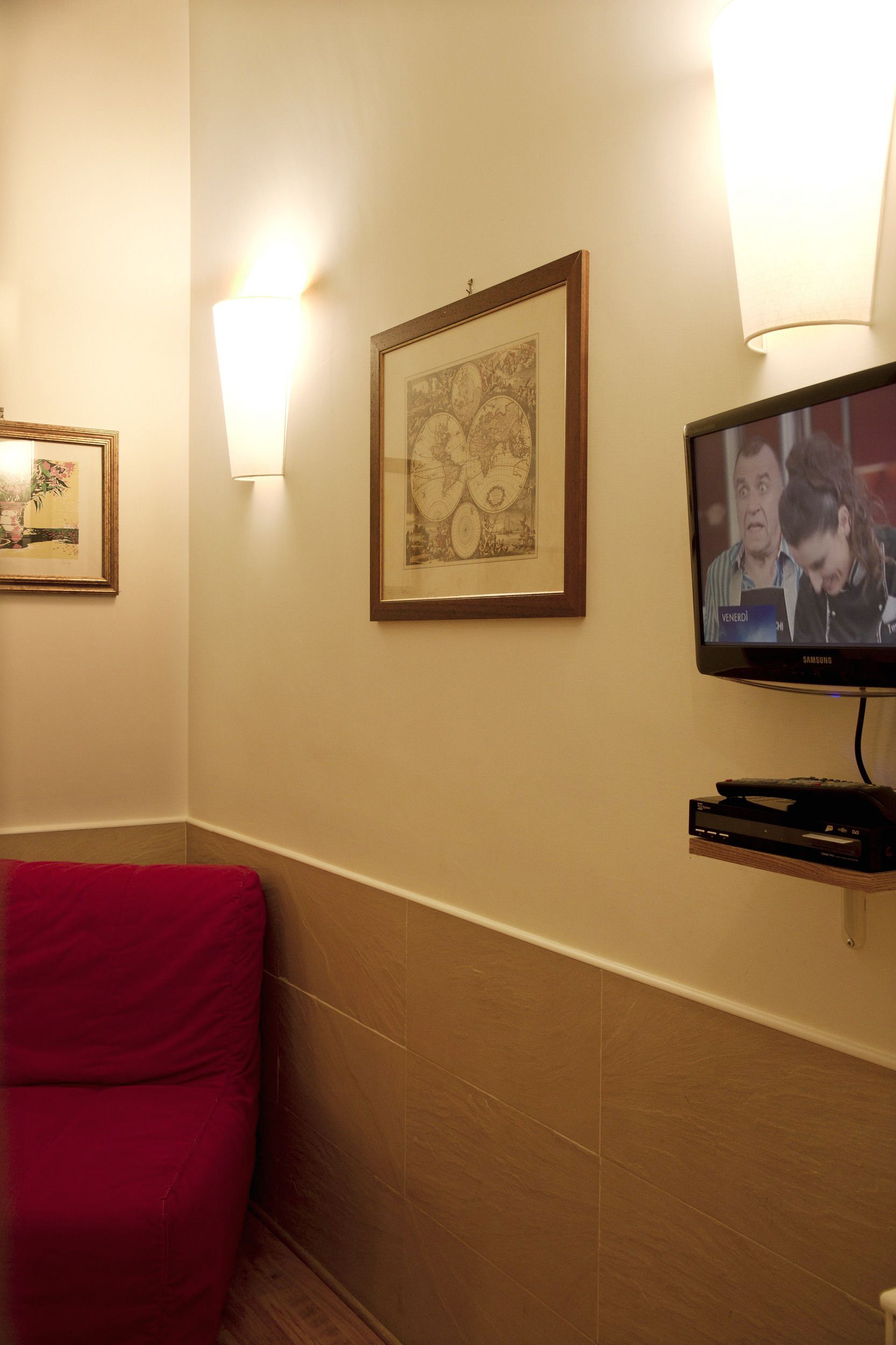 A hotel room with a red chair, a framed artwork, a television, and a wall-mounted light fixture.