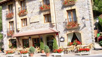 Un edificio de piedra con techo de tejas, múltiples balcones con macetas de flores, mesas y sillas al aire libre y un letrero que indica que es un hotel.