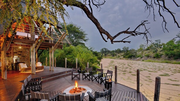 Een terras buiten met een vuurkuil, omringd door stoelen, met uitzicht op een zandstrand en een stenen gebouw met rieten dak.