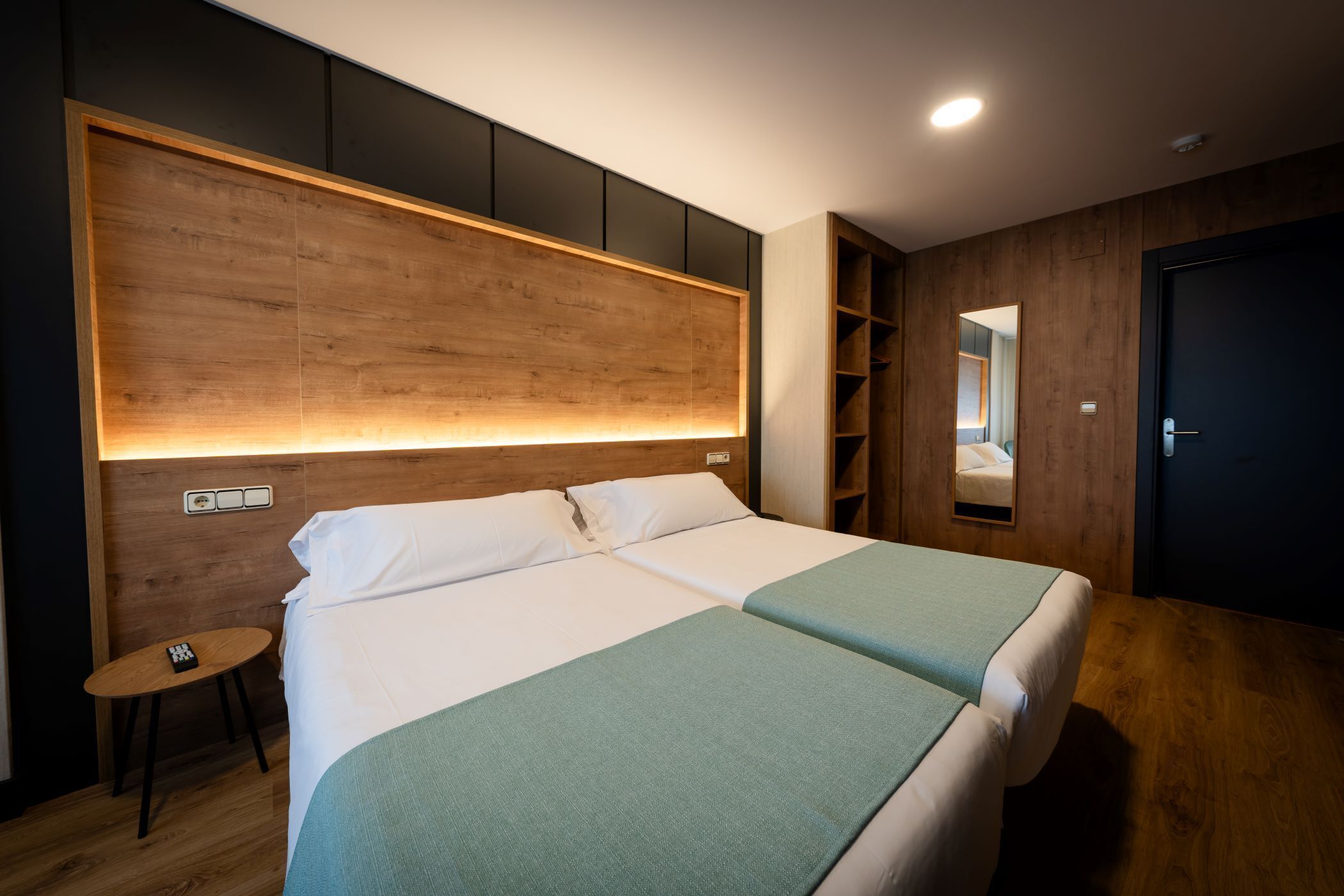 Una habitación de hotel moderna con una cama grande, cabecera de madera, mesitas de noche y una estantería empotrada.