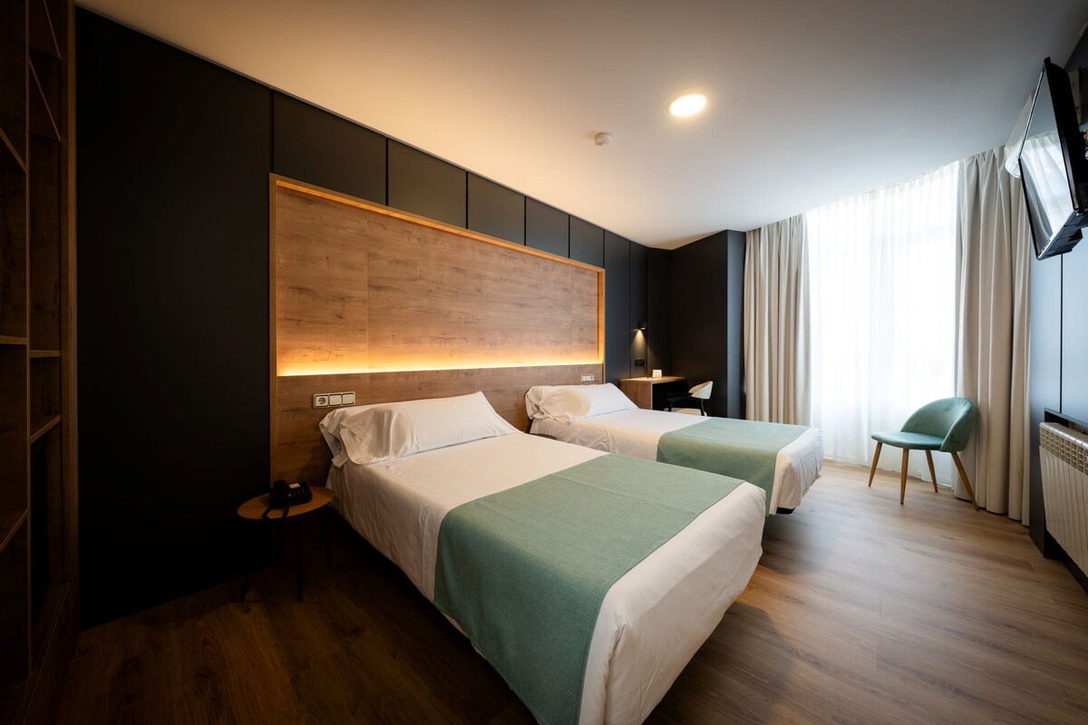 Une chambre d’hôtel avec deux lits, une tête de lit en bois, une table de chevet, une chaise et un téléviseur fixé au mur.