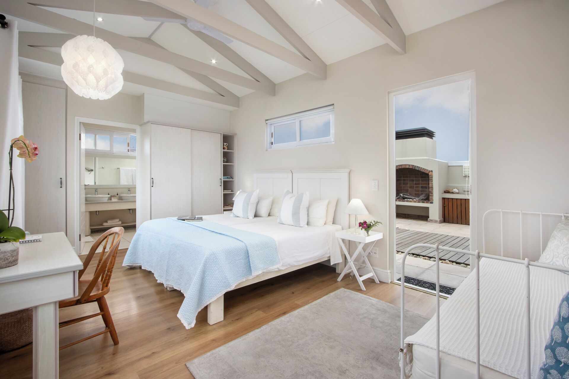 Une chambre moderne avec un grand lit, un bureau, une chaise, une petite table avec une lampe, et une vue sur un patio extérieur doté d’une cheminée.