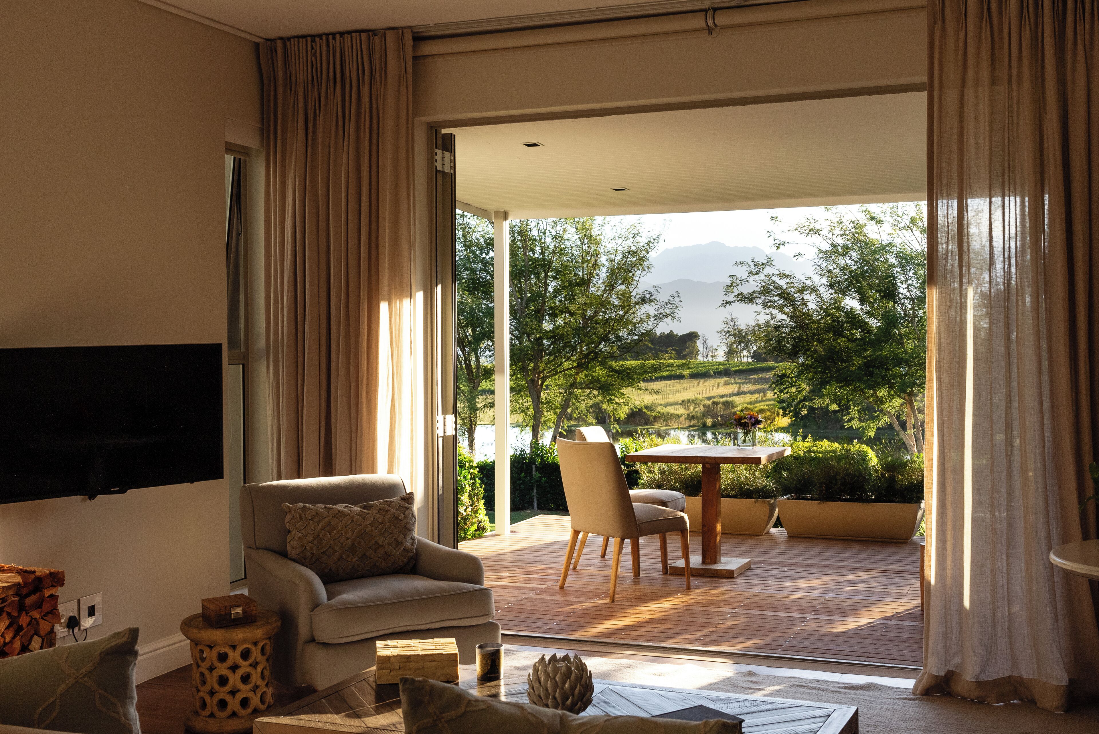 A modern living room with a sofa, a coffee table, and a TV mounted on the wall. There is a sliding glass door leading to an outdoor patio with a table and chairs.