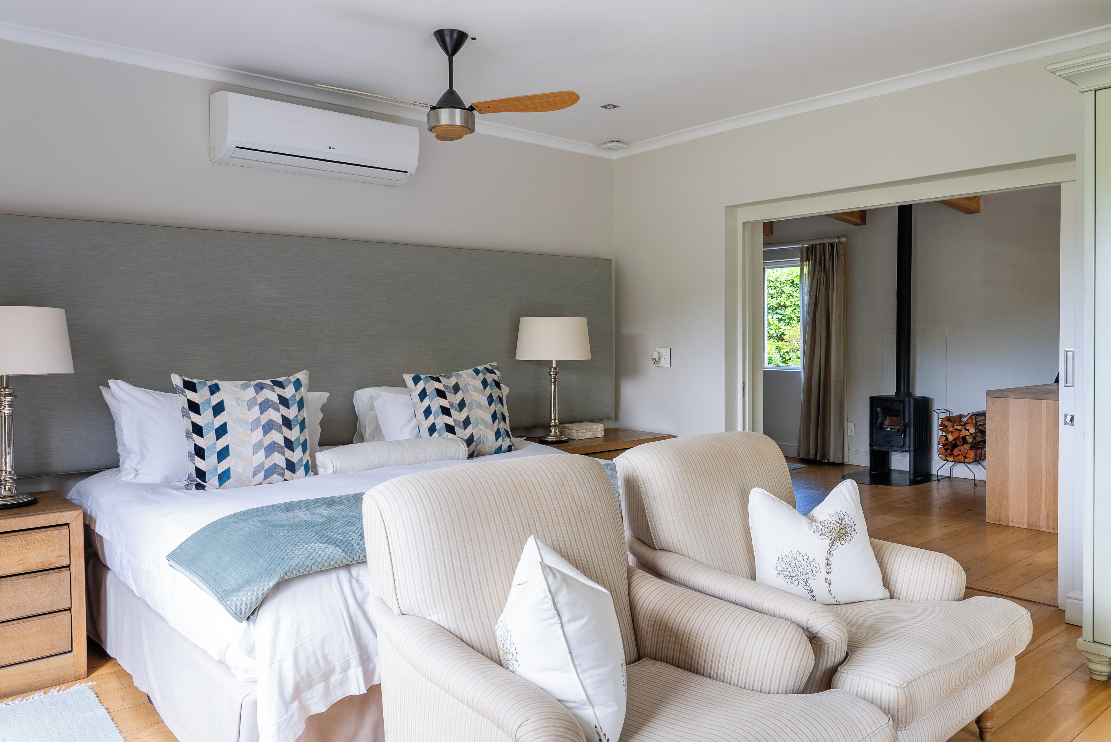 A bedroom with a bed, two bedside tables with lamps, a ceiling fan, and a view of a living area with a fireplace.