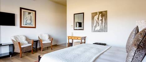 A bedroom with a large bed, two wicker chairs, a small wooden desk, and artwork on the walls.