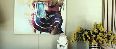 A painting of a woman holding a blue object, a vase with yellow flowers, and a white sculpture on a dark wooden table.
