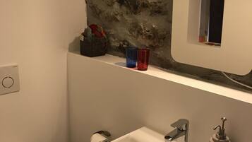 Une salle de bain avec un lavabo blanc, un miroir et des toilettes. Le mur derrière le lavabo est texturé et une petite étagère y est fixée, sur laquelle se trouvent un sac et deux tasses.
