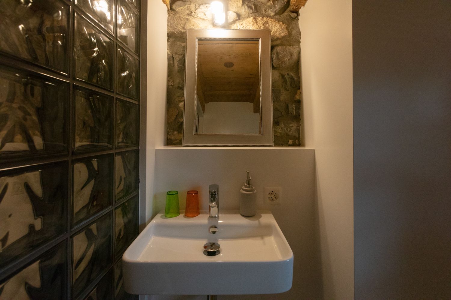 A bathroom with a white sink, a mirror above it, and a stone wall behind the mirror. There are three colorful bottles on the sink's edge.