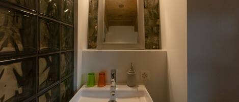 A bathroom with a white sink, a mirror above it, and a stone wall behind the mirror. There are three colorful bottles on the sink's edge.