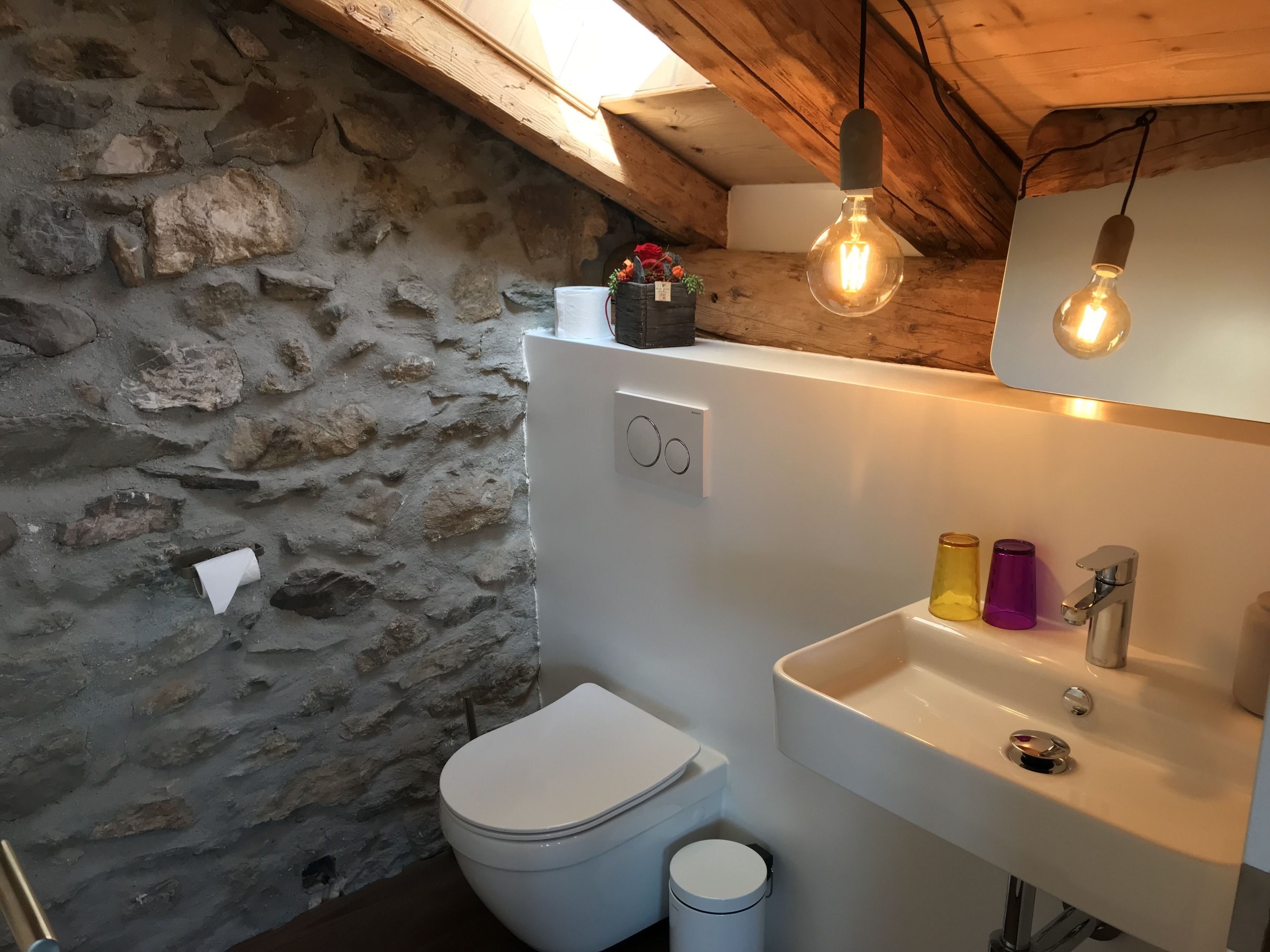 Une salle de bain avec un mur en pierre, des toilettes blanches, un lavabo avec un miroir et deux lampes suspendues.