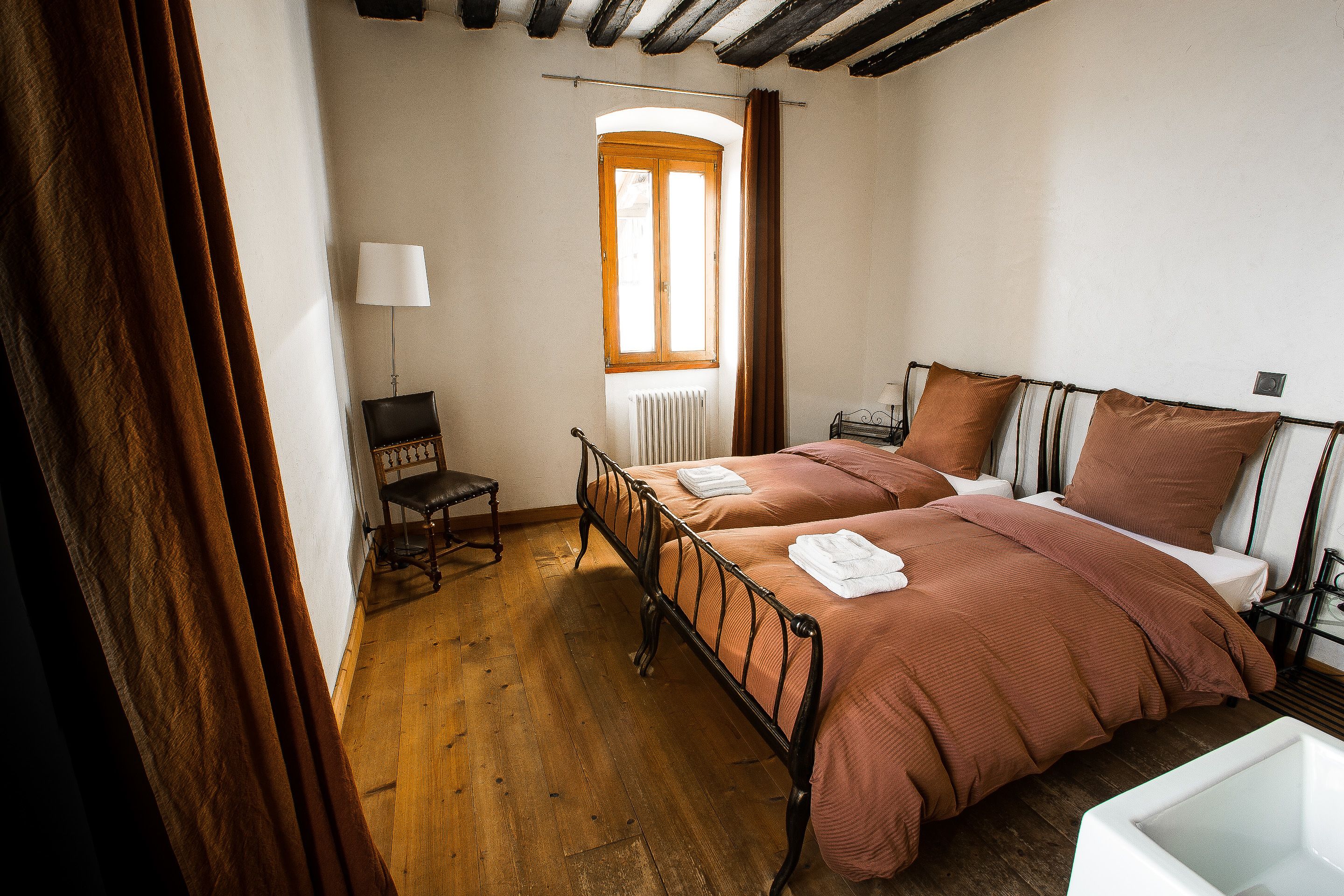 Un dormitorio con suelo de madera, una cama con colcha marrón, una silla, una lámpara y una ventana con cortinas.