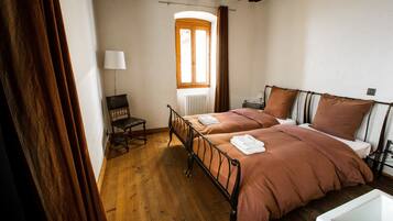 Un dormitorio con suelo de madera, una cama con colcha marrón, una silla, una lámpara y una ventana con cortinas.