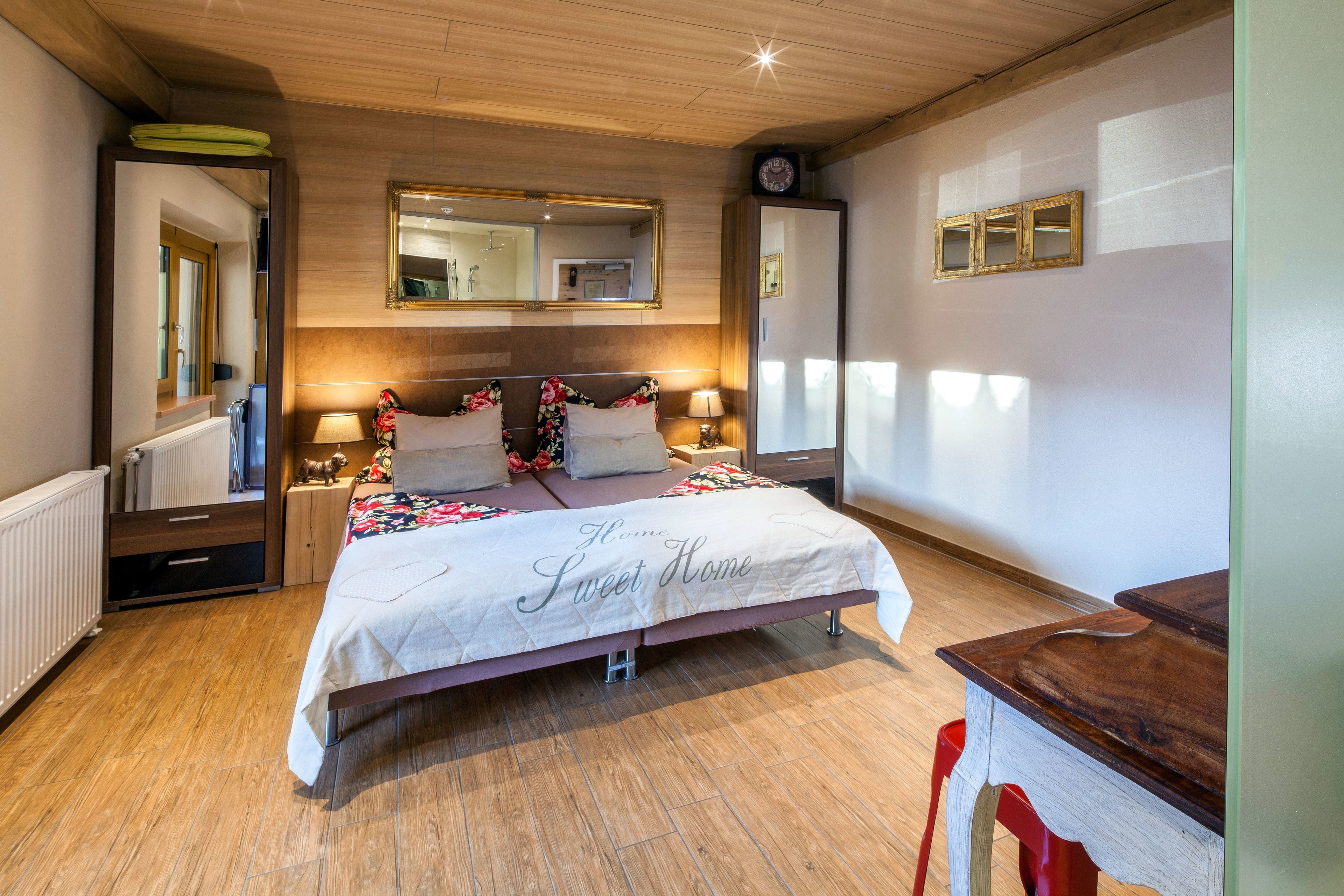 A bedroom with a wooden ceiling, a large bed with a floral bedspread, a mirror, a dresser, and a nightstand.