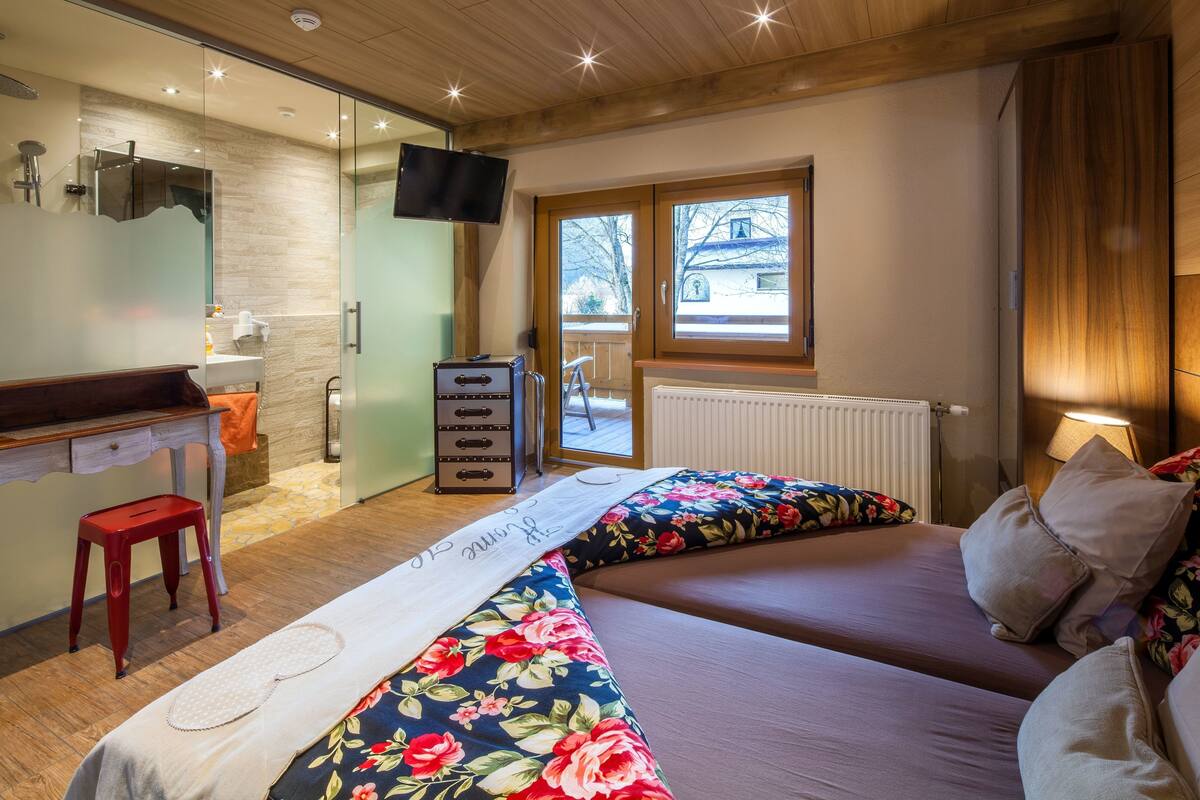 A bedroom with two beds, a desk, a chair, a TV, and a window with a view of the outside.