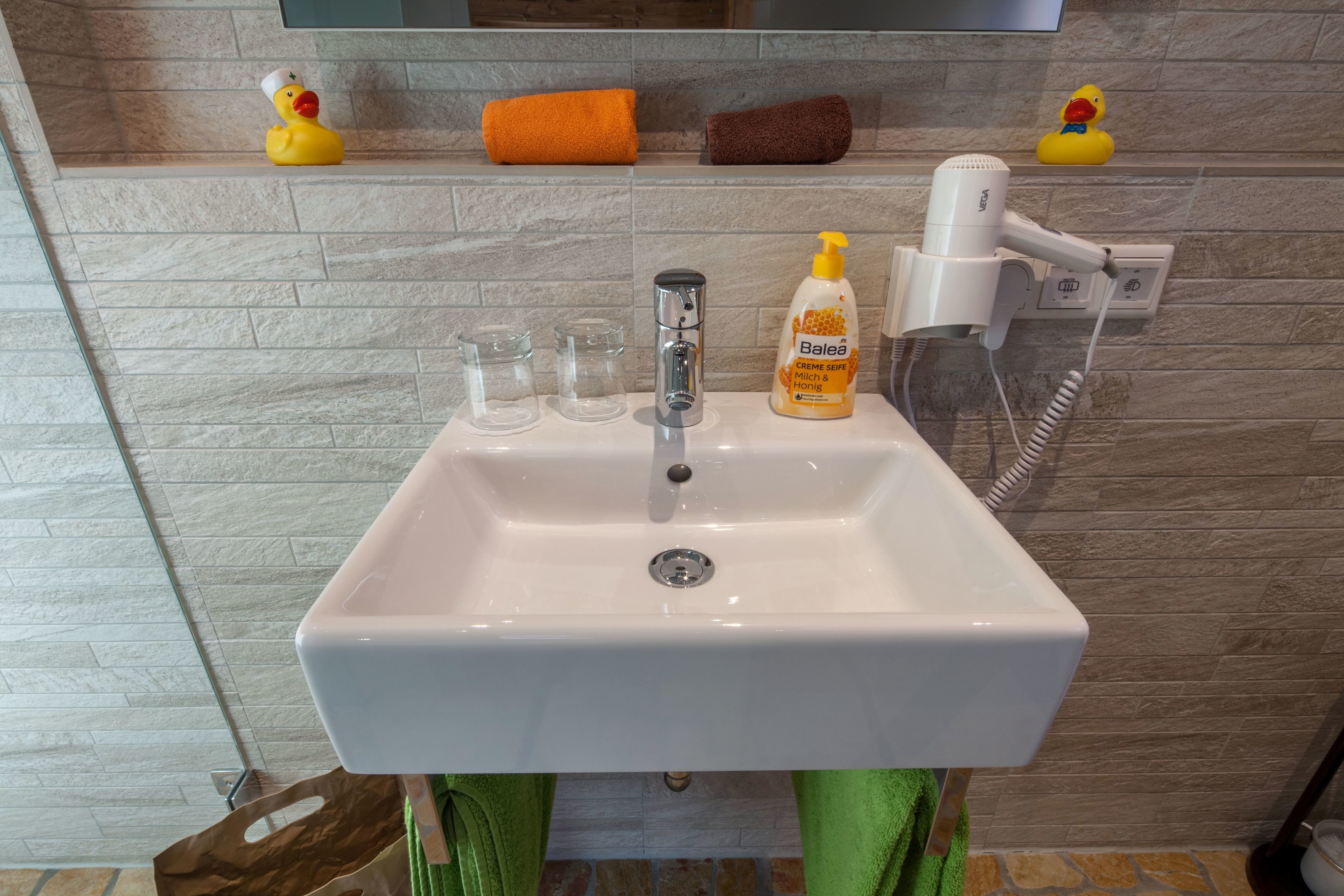 A bathroom with a white sink, a hairdryer, and a bottle of shampoo. There are rubber ducks on the wall and a towel rack with towels.