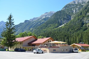 A mountain lodge with a red-roofed building, a parking area, and a view of the surrounding forested mountains. - Gasthaus - Pension Reiterklause (Leutasch)
