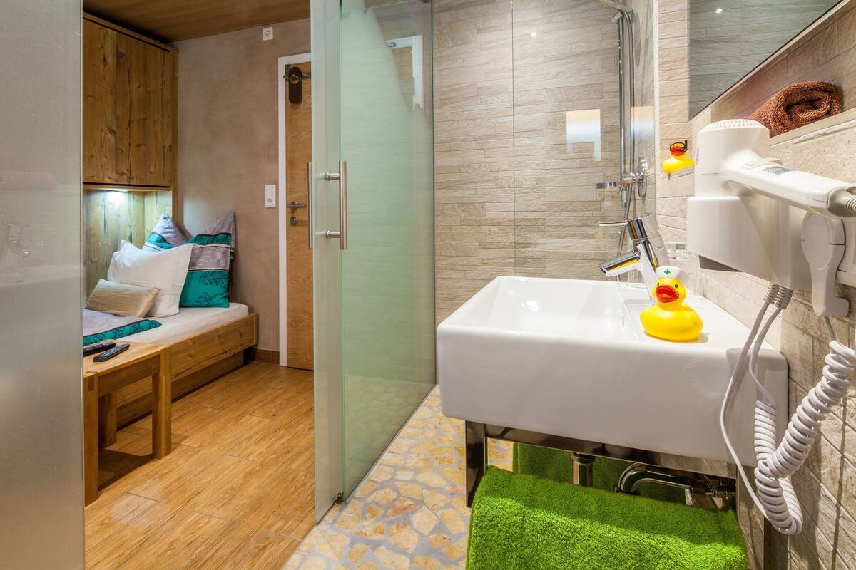 A compact room with a wooden bed, a glass shower enclosure, a white sink with a rubber duck, and a green towel.
