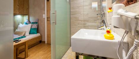 A compact room with a wooden bed, a glass shower enclosure, a white sink with a rubber duck, and a green towel.
